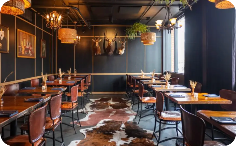 Warmly lit restaurant dining room with wooden tables, leather chairs, rattan pendants, and a cowhide rug